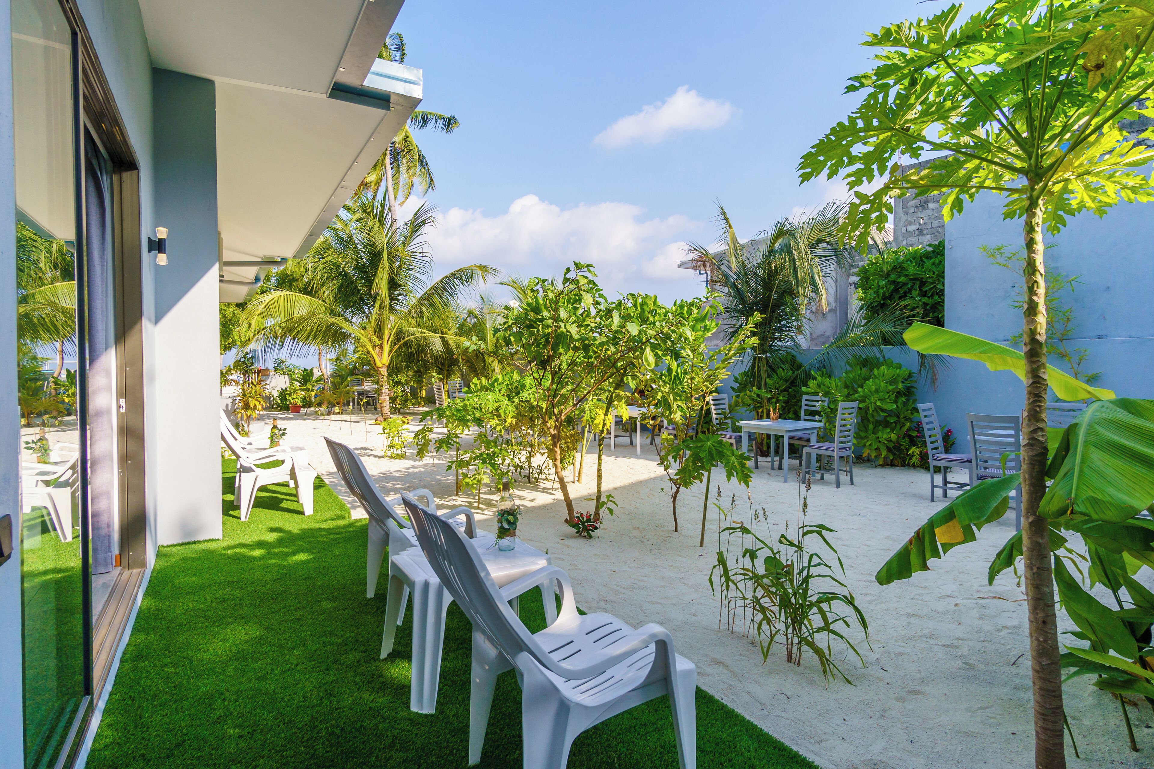Un patio con sillas y mesas de plástico blanco, rodeado de césped artificial y arena, con palmeras y otras plantas tropicales.