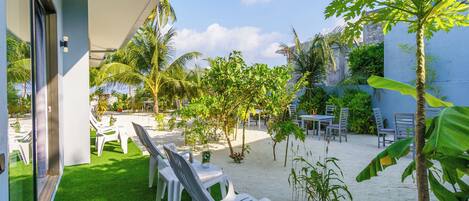 Un patio con sillas y mesas de plástico blanco, rodeado de césped artificial y arena, con palmeras y otras plantas tropicales.