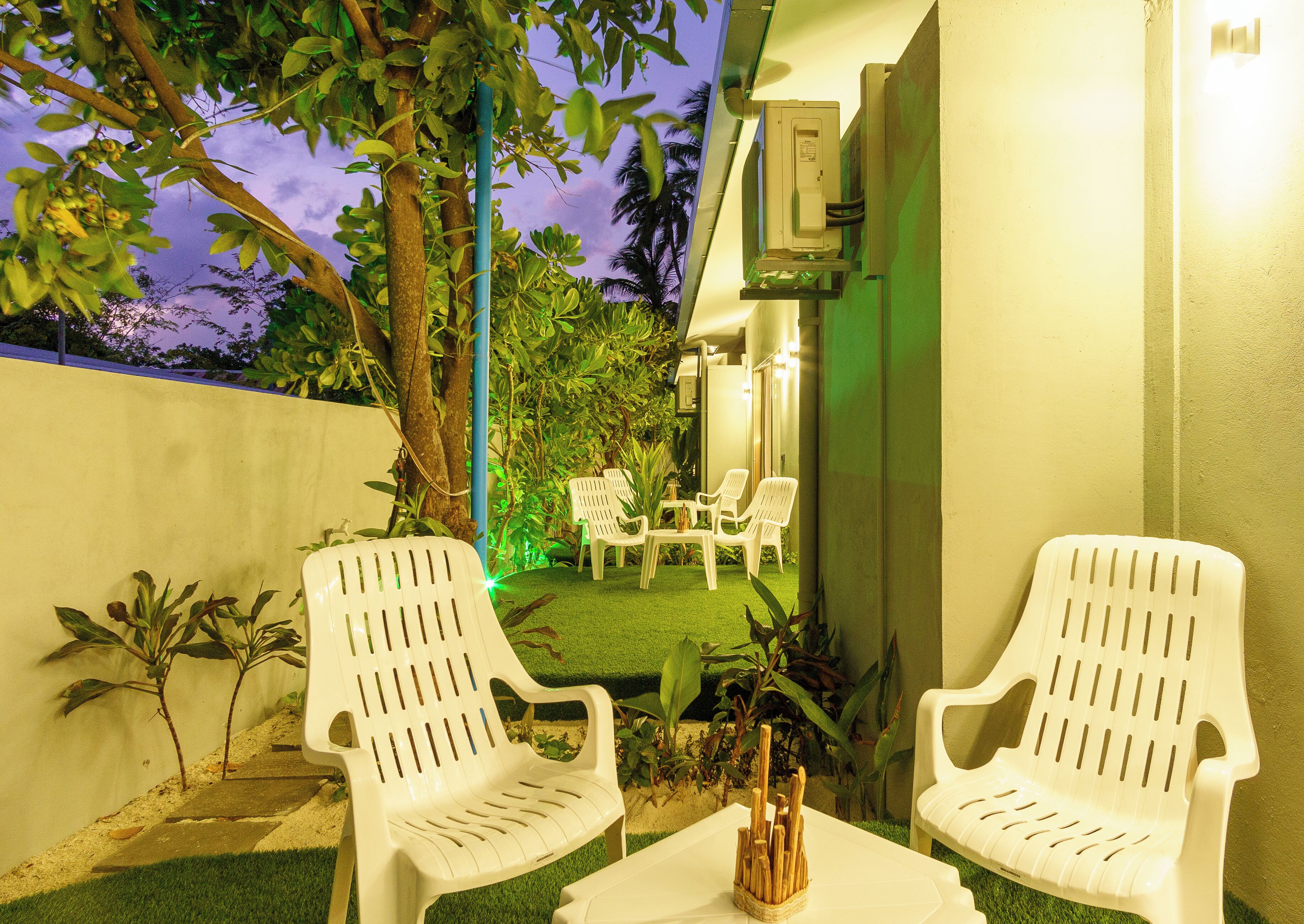 Two white plastic chairs with a small table in between, a green artificial lawn, and a building with a light fixture in the background.