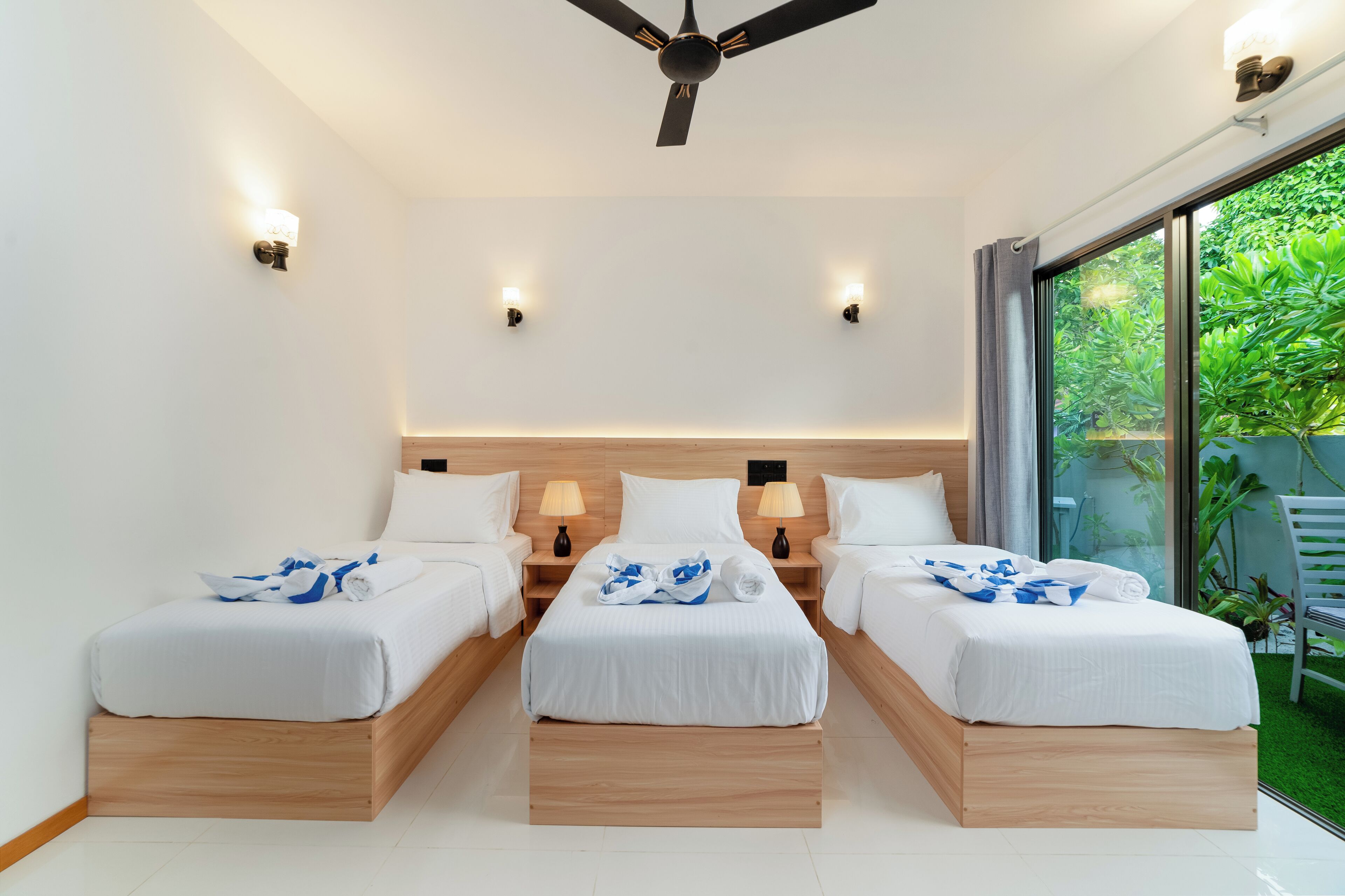 Habitación de hotel con tres camas individuales, ventilador de techo, lámparas de noche y un amplio ventanal corredero a un jardín.