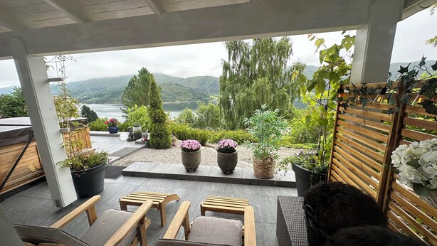 Un espace extérieur aménagé, abrité et doté de chaises longues en bois et de plantes en pot, offrant une vue sur un lac et des montagnes.