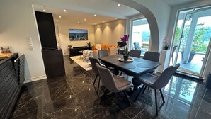 Un espace repas moderne avec un sol en marbre, une table à manger et des chaises, un coin cuisine doté d’un réfrigérateur noir et une grande fenêtre donnant sur un paysage verdoyant.
