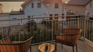 A balcony with wicker chairs and a small table, overlooking a row of houses and a body of water during sunset.