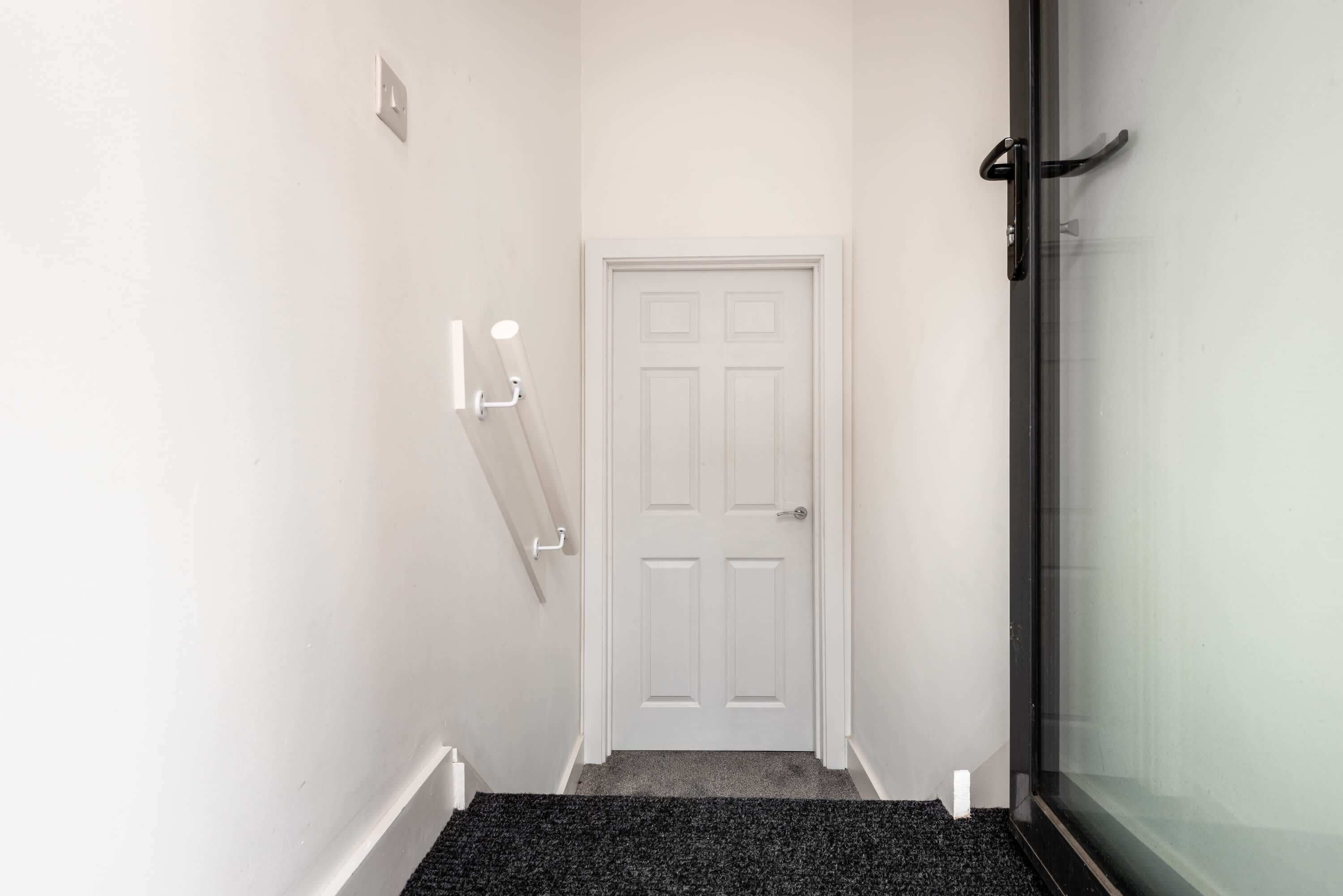 A narrow hallway with a white door, a coat rack, and a glass door leading to a room.