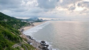 A coastal landscape with a beach, rocky shoreline, and distant buildings. - Weifudun Holiday Apartment (Hailing Island Poly Silver Beach) (Yangjiang)
