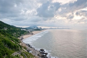 A coastal landscape with a beach, rocky shoreline, and distant buildings. - Weifudun Holiday Apartment (Hailing Island Poly Silver Beach) (Yangjiang)