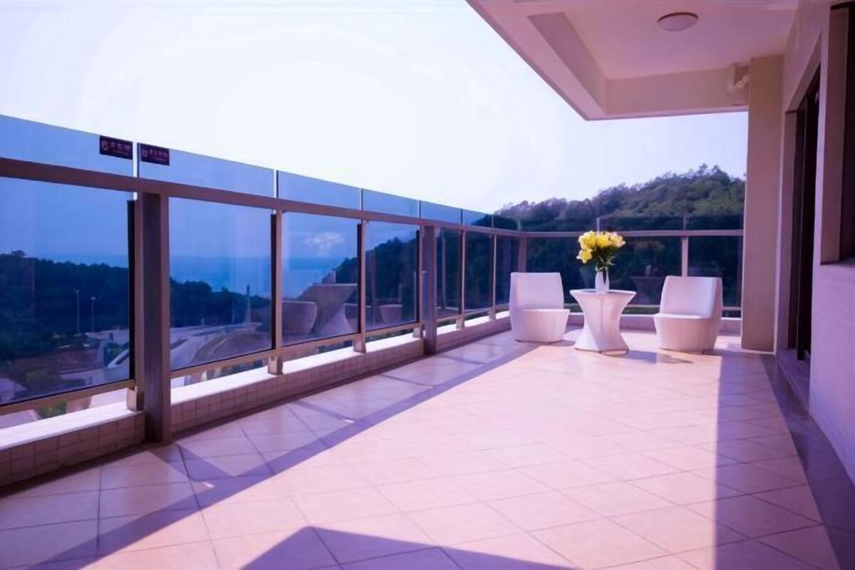 Un balcón con vistas a un paisaje montañoso, sillas blancas y una mesa con flores.