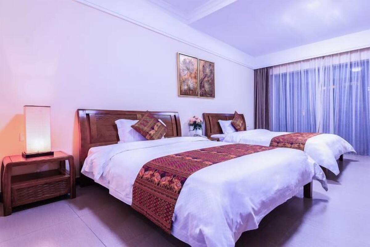 Habitación de hotel con dos camas, una mesita de noche, una lámpara, un cuadro en la pared y un ventanal con cortinas.