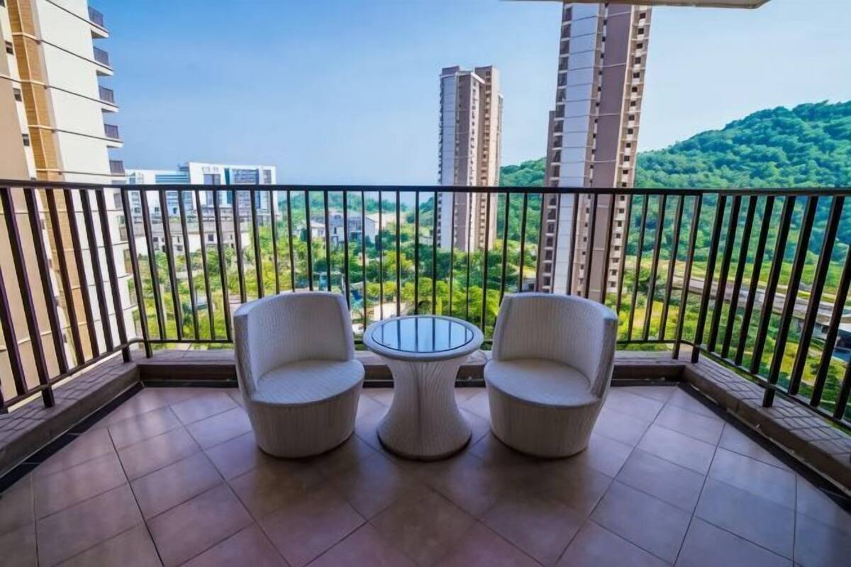 Un balcón con dos sillas blancas y una mesita redonda, con vistas a un paisaje urbano con edificios altos y zonas verdes.