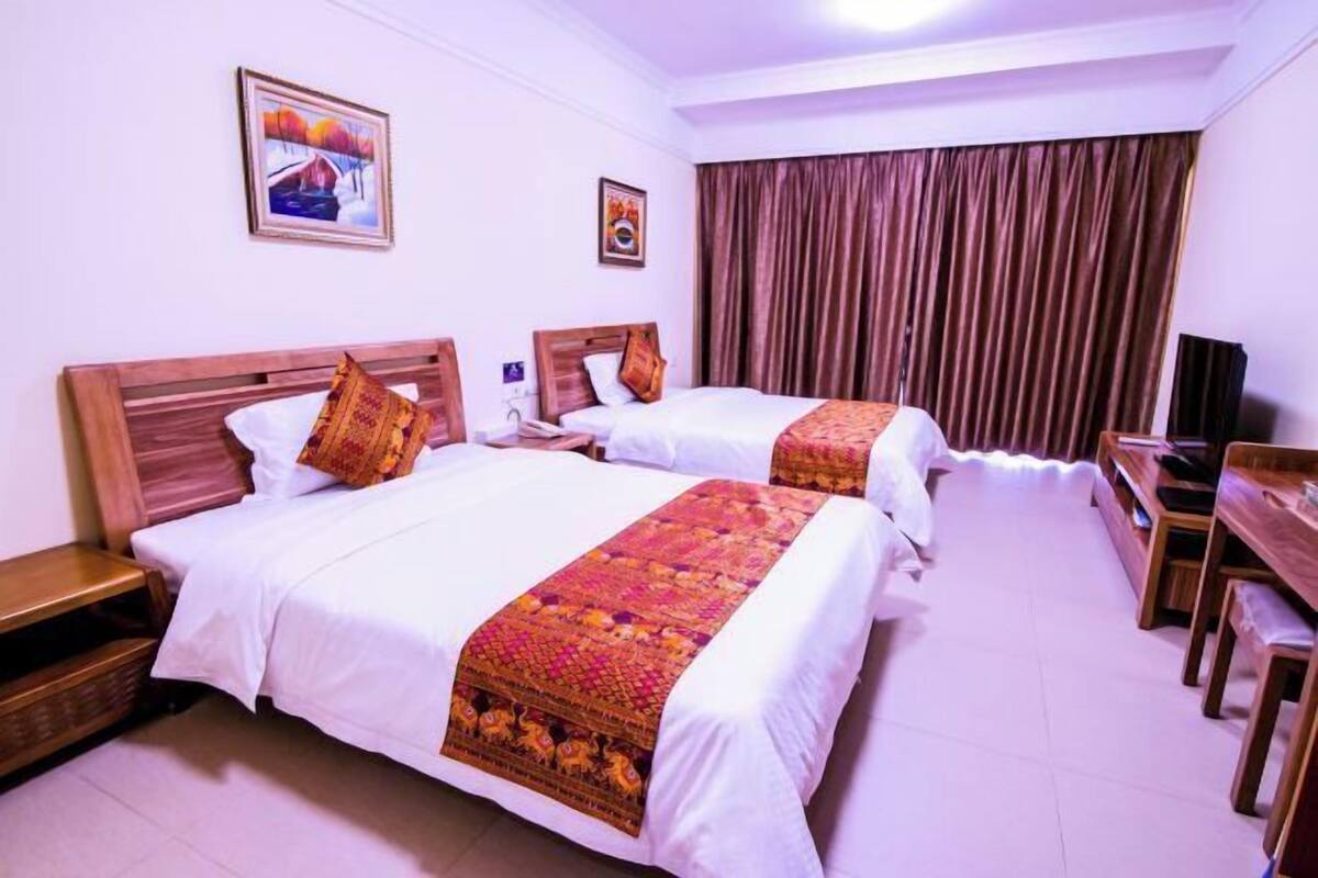 Habitación de hotel con dos camas, cabeceros de madera, ropa de cama blanca, una manta estampada, un televisor y una cortina.