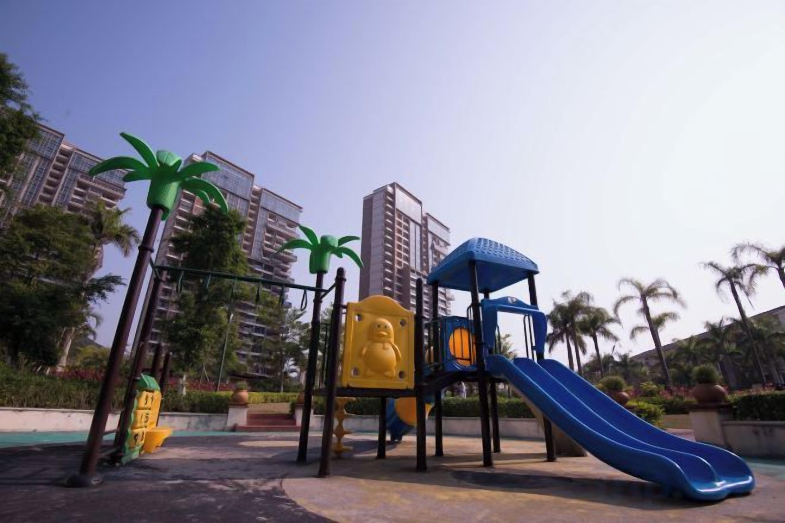 A playground with a slide and climbing structures, surrounded by palm trees and residential buildings.