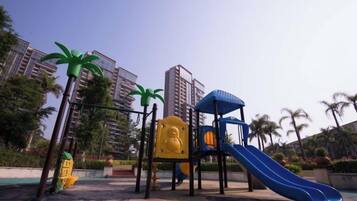 A playground with a slide and climbing structures, surrounded by palm trees and residential buildings.