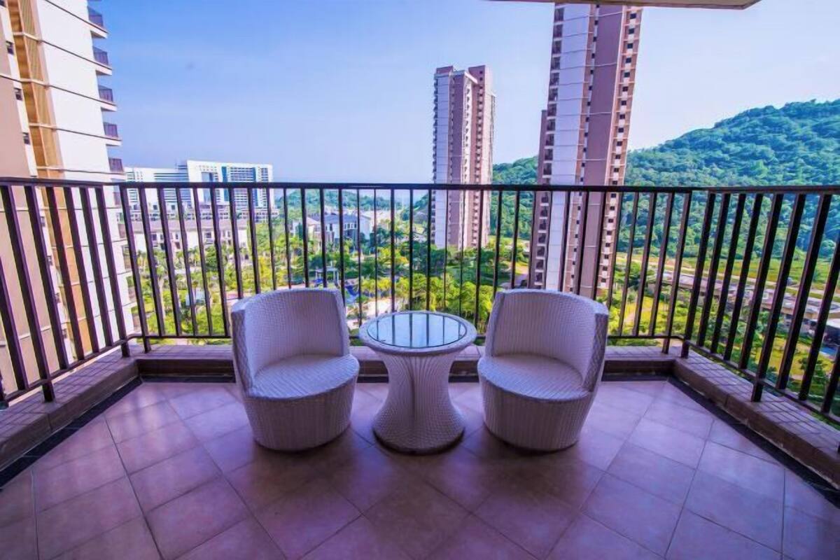 Un balcón con dos sillas de mimbre y una mesa de vidrio, con vistas a un paisaje urbano con edificios altos y una colina verde al fondo.