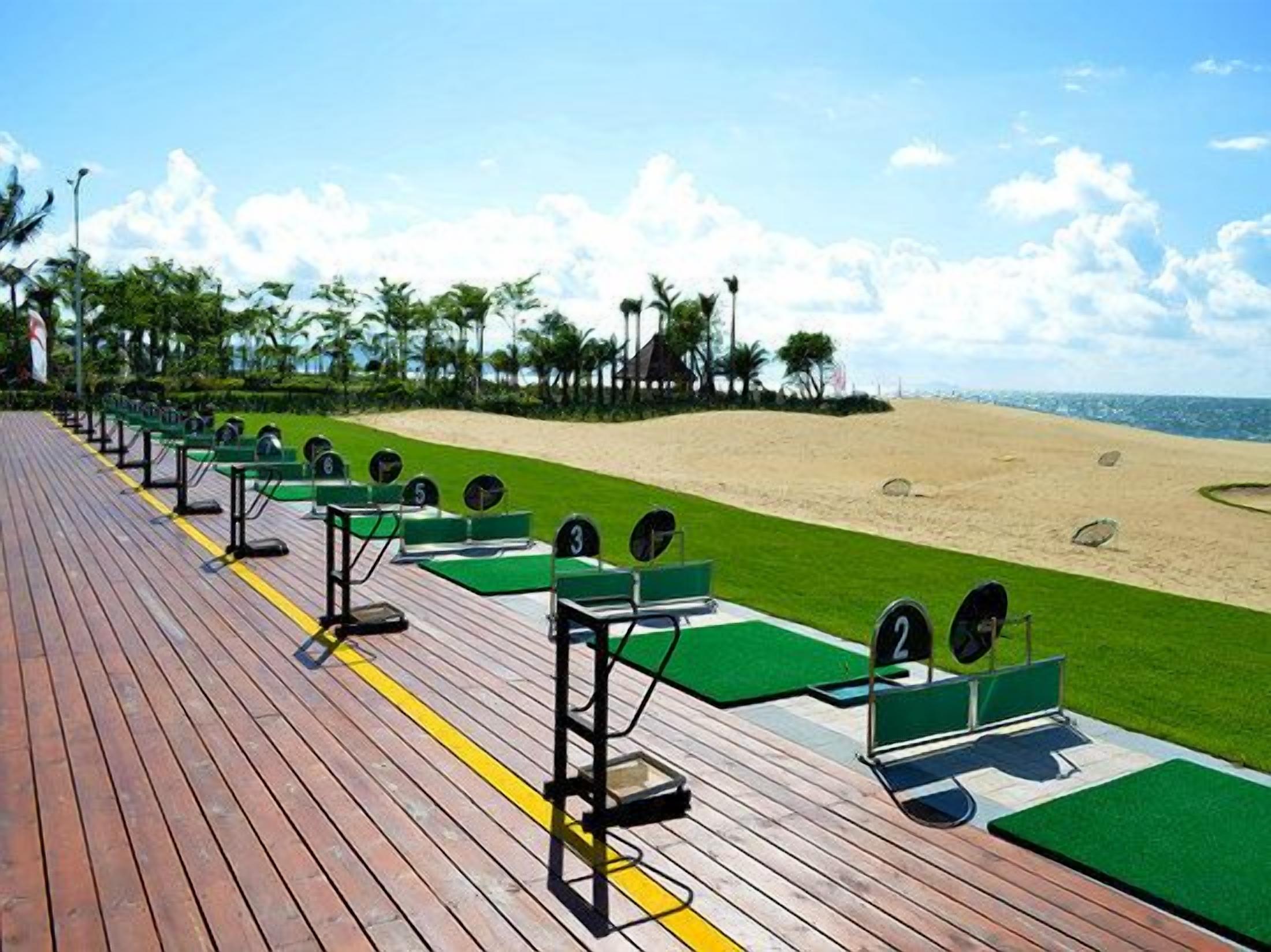 A wooden deck with exercise equipment and green mats, overlooking a sandy beach with palm trees.