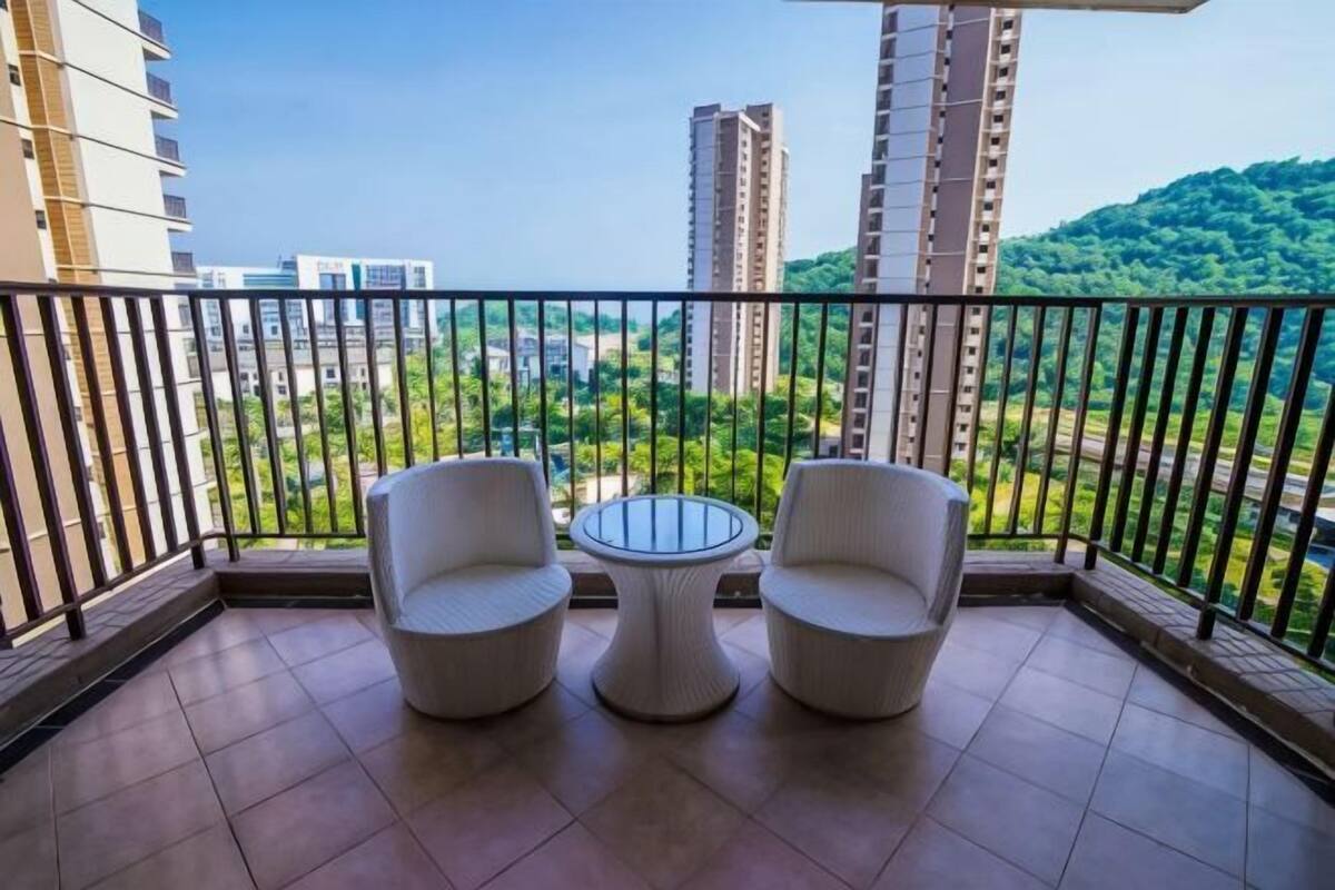 Un balcón con dos sillas blancas y una mesita pequeña, con vistas a un paisaje urbano con edificios altos y zonas verdes.