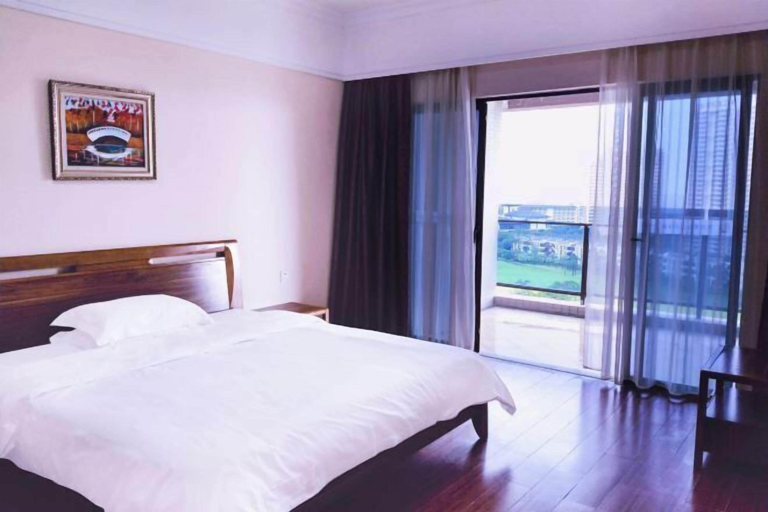 A hotel room with a large bed, a wooden headboard, a nightstand, a framed picture on the wall, and a balcony with curtains.
