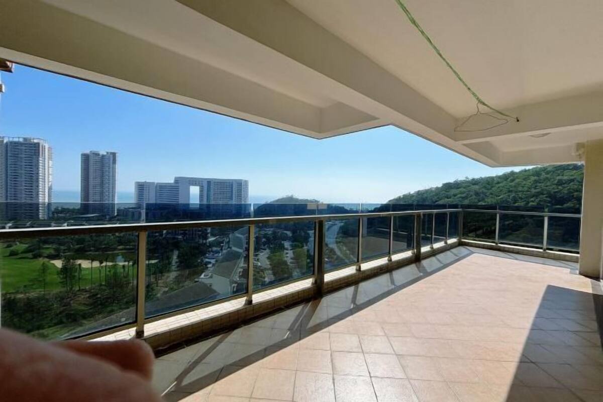Un balcón con barandilla de vidrio, suelo de baldosas y vistas a edificios y vegetación.