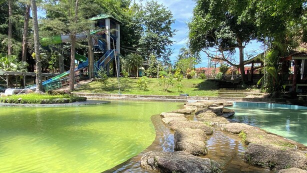 Uma piscina translúcida com pedras em primeiro plano, um parquinho com escorregadores e um edifício com telhado verde ao fundo.