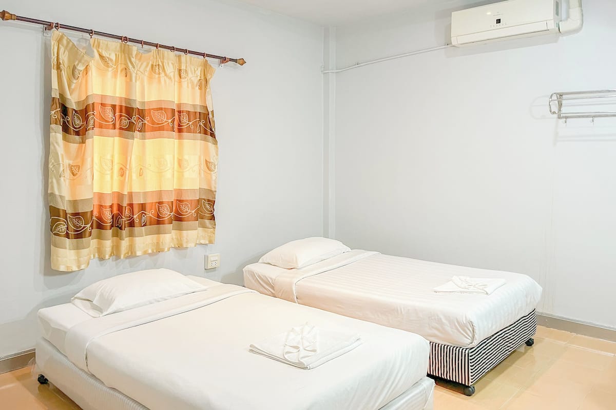 Habitación de hotel con dos camas individuales, cortina estampada con motivos de hojas, unidad de aire acondicionado blanca y suelo de baldosas.