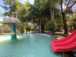 A water feature with a mushroom-shaped structure, a red slide, and a pool surrounded by trees.