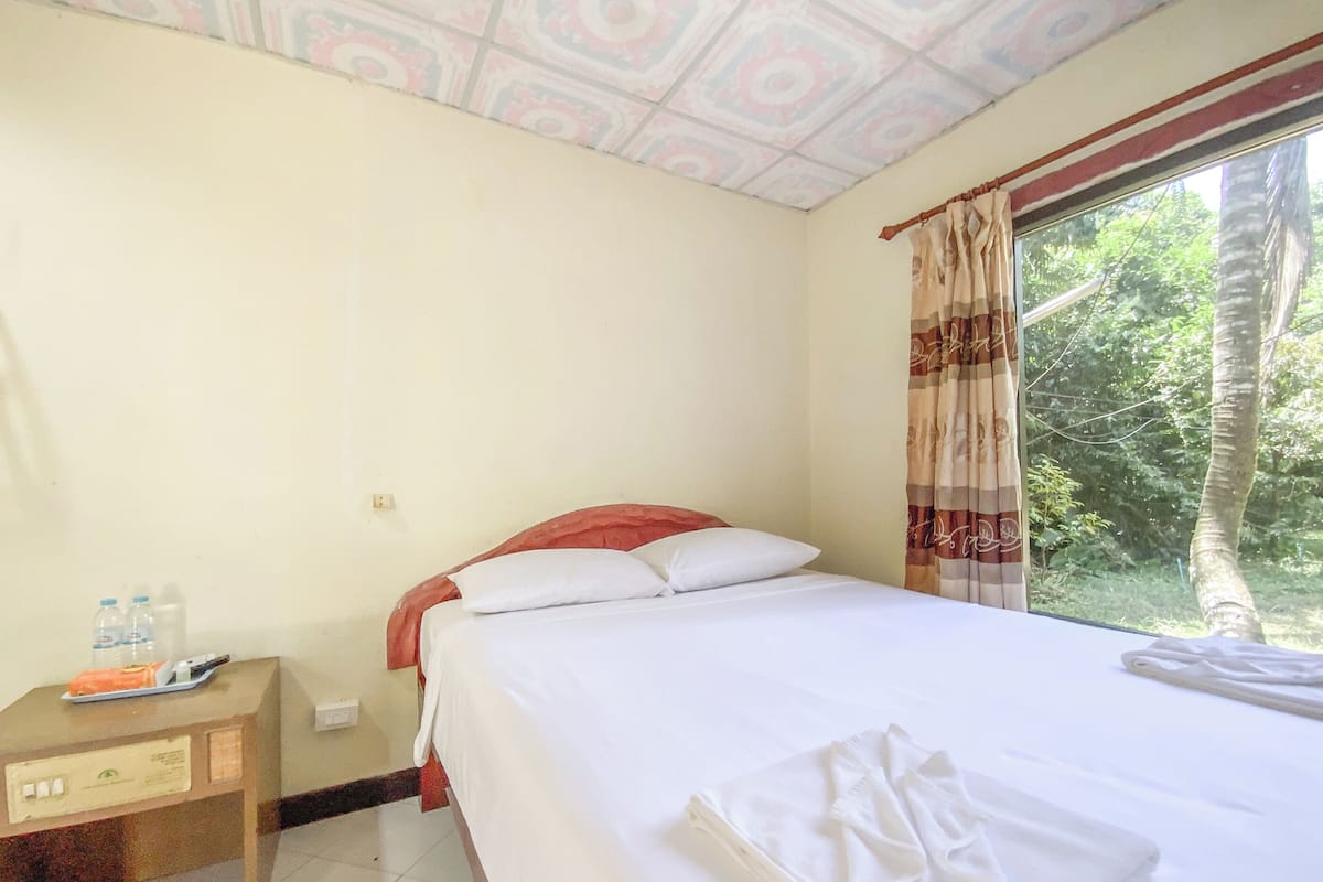 Habitación de hotel con cama, mesita de noche, ventana con cortinas y vista a árboles.