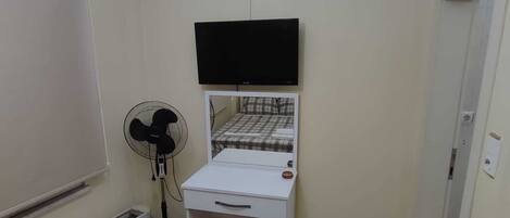 A bedroom with a bed, a desk, a chair, a standing fan, and a heater.