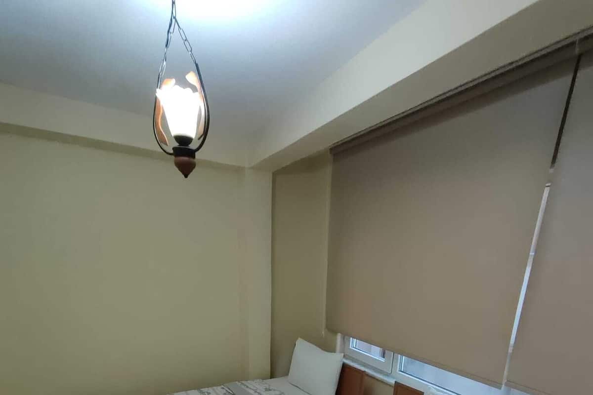 A bedroom with a large bed, a window with blinds, and a hanging light fixture.