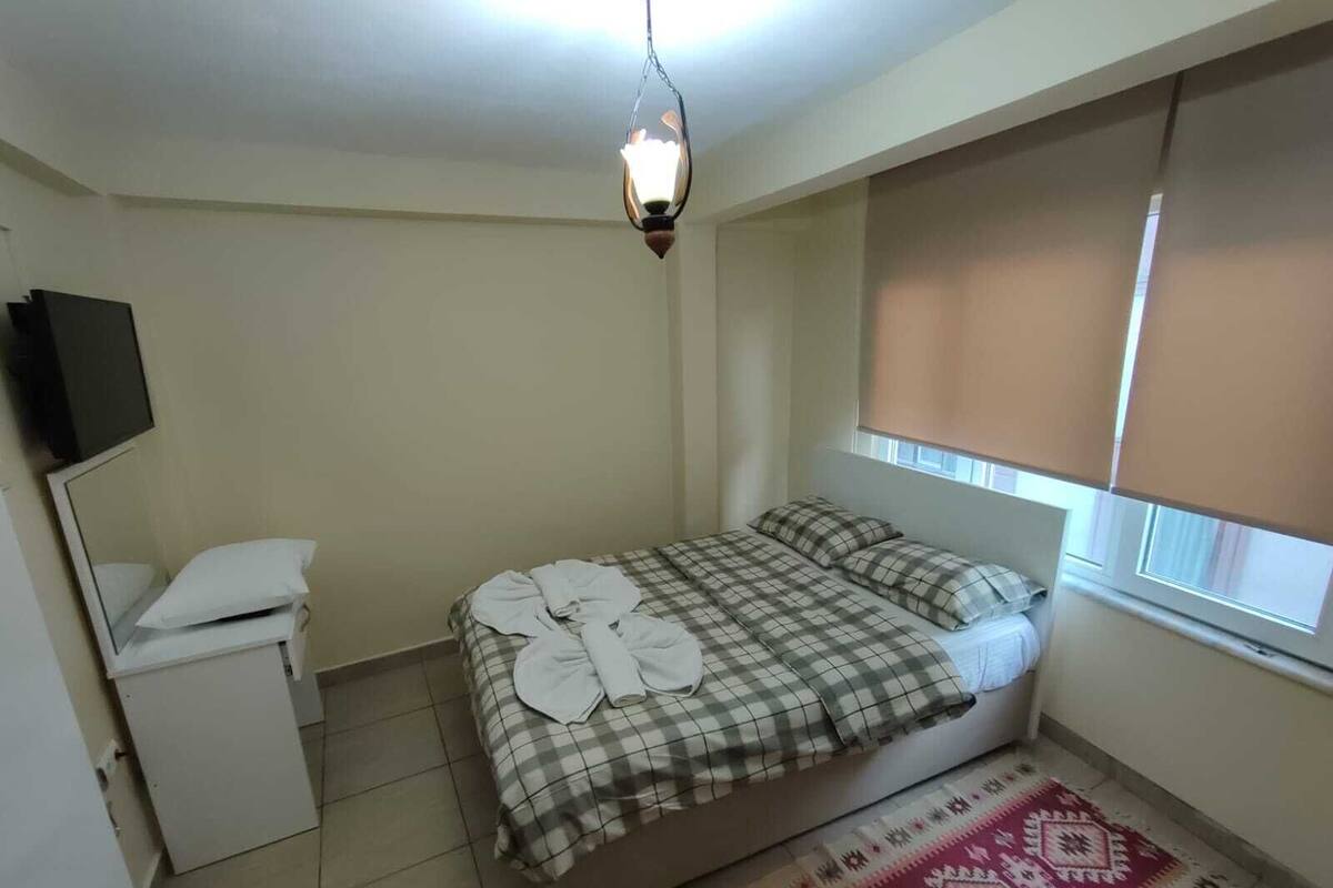 A small, clean room with a bed, a bedside table, a mirror, and a window with blinds.