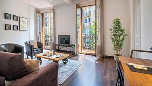 A modern living room with a sofa, armchairs, a coffee table, and a TV. There is a large window with a view of buildings and a plant. - Habitat Apartments Barcelona Balconies (Barcelona)