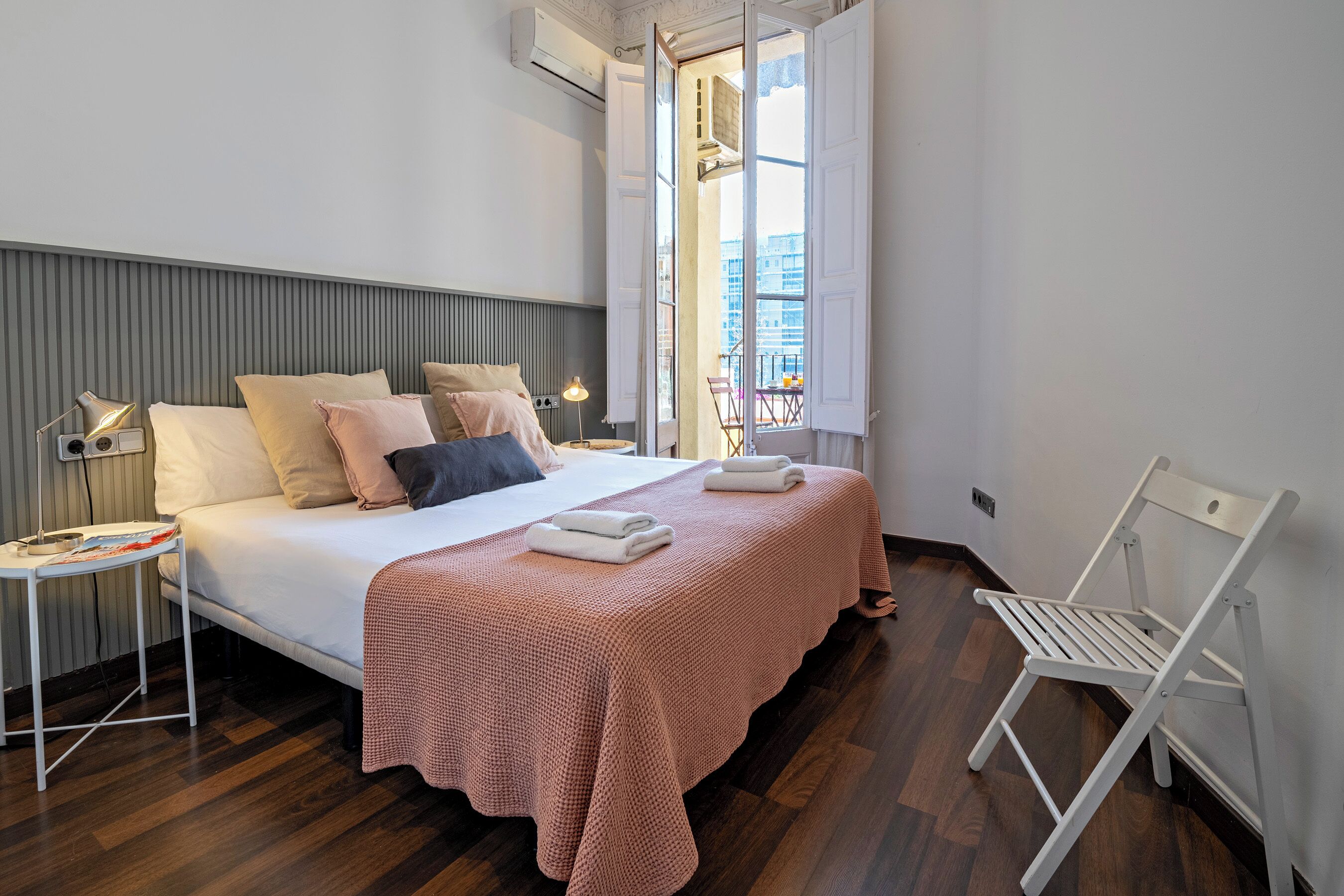 A modern bedroom with a bed, bedside tables, a chair, and a window with a view of buildings.