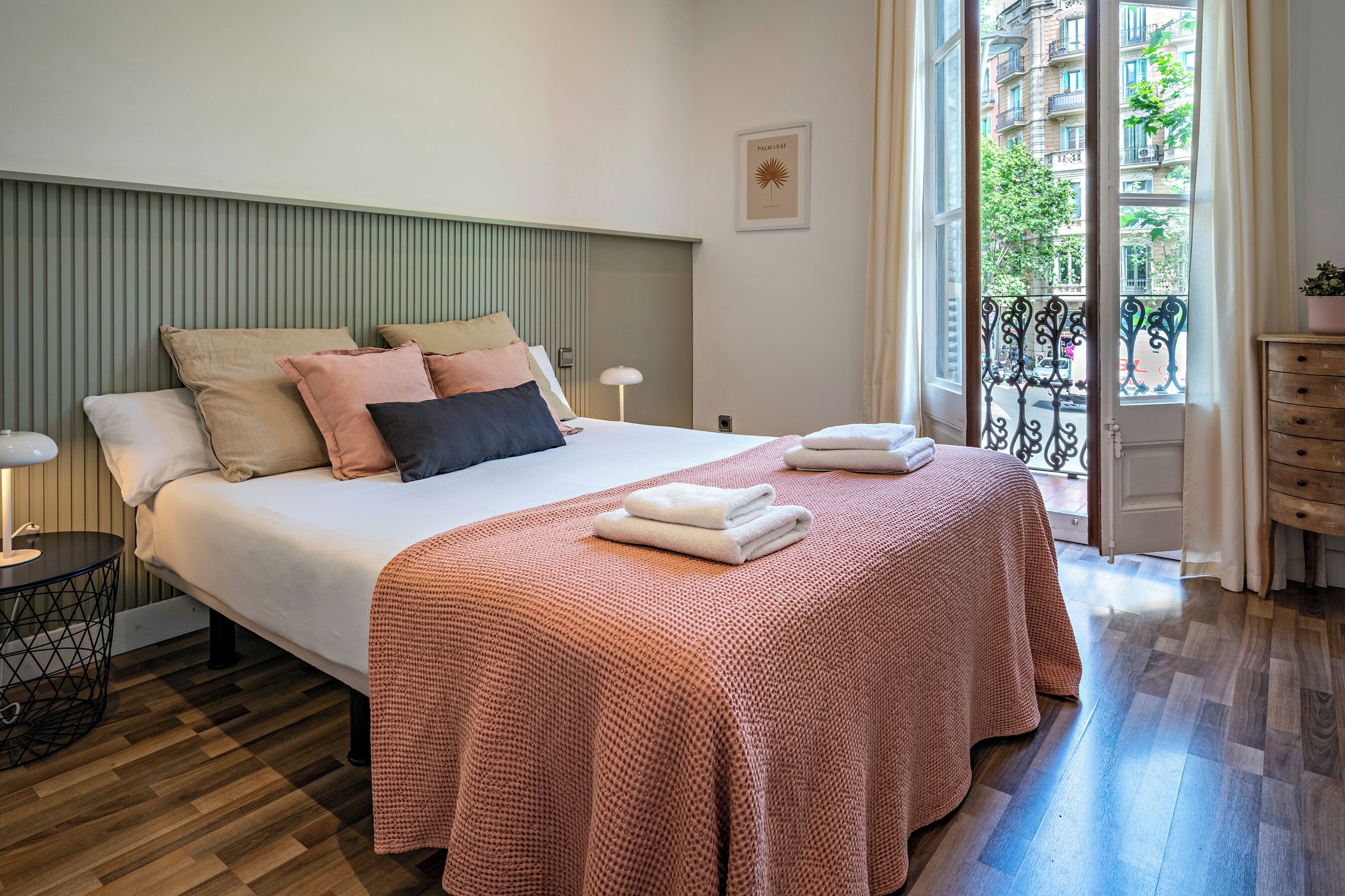 A neatly arranged bedroom with a bed, pillows, a bedside table with a lamp, a dresser, and a view of the outside balcony.
