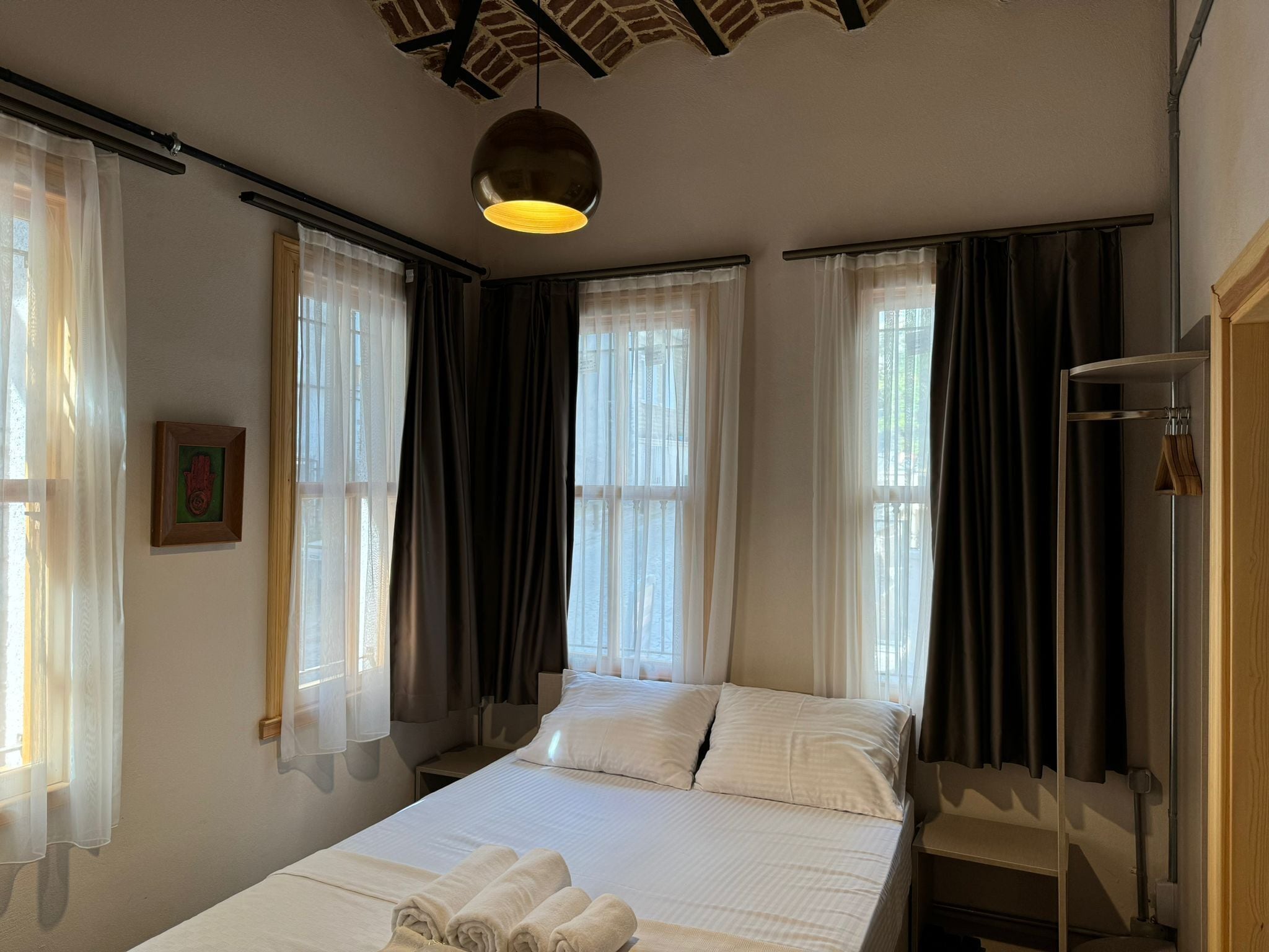 A hotel room with a bed, two windows with curtains, a hanging light fixture, and a wooden door.