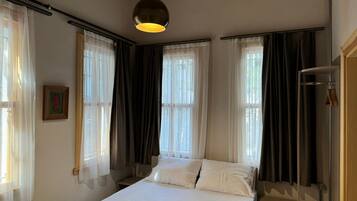 A hotel room with a bed, two windows with curtains, a hanging light fixture, and a wooden door.