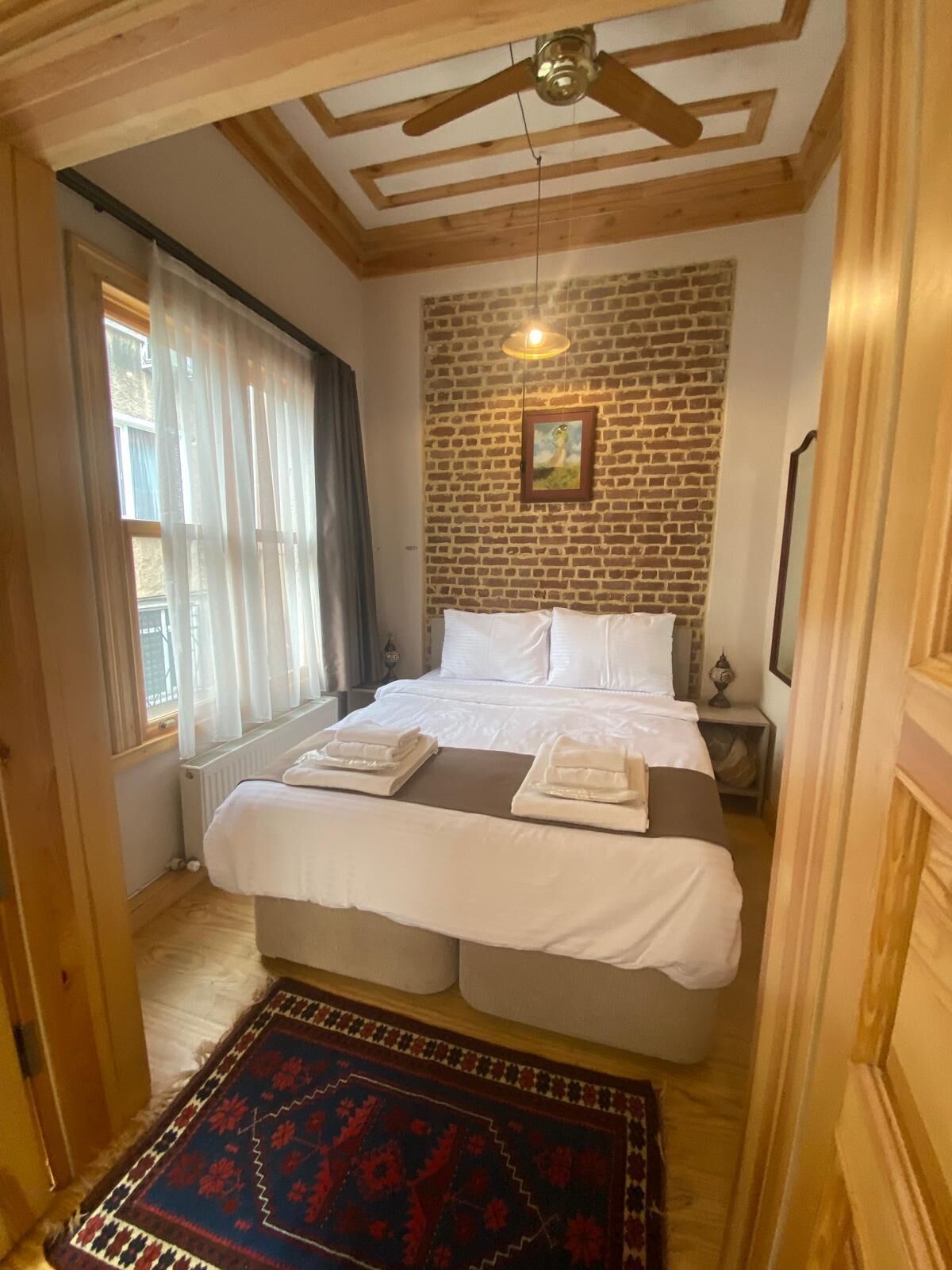 A cozy bedroom with a brick wall, a wooden ceiling, a bed with white linens, and a patterned rug.