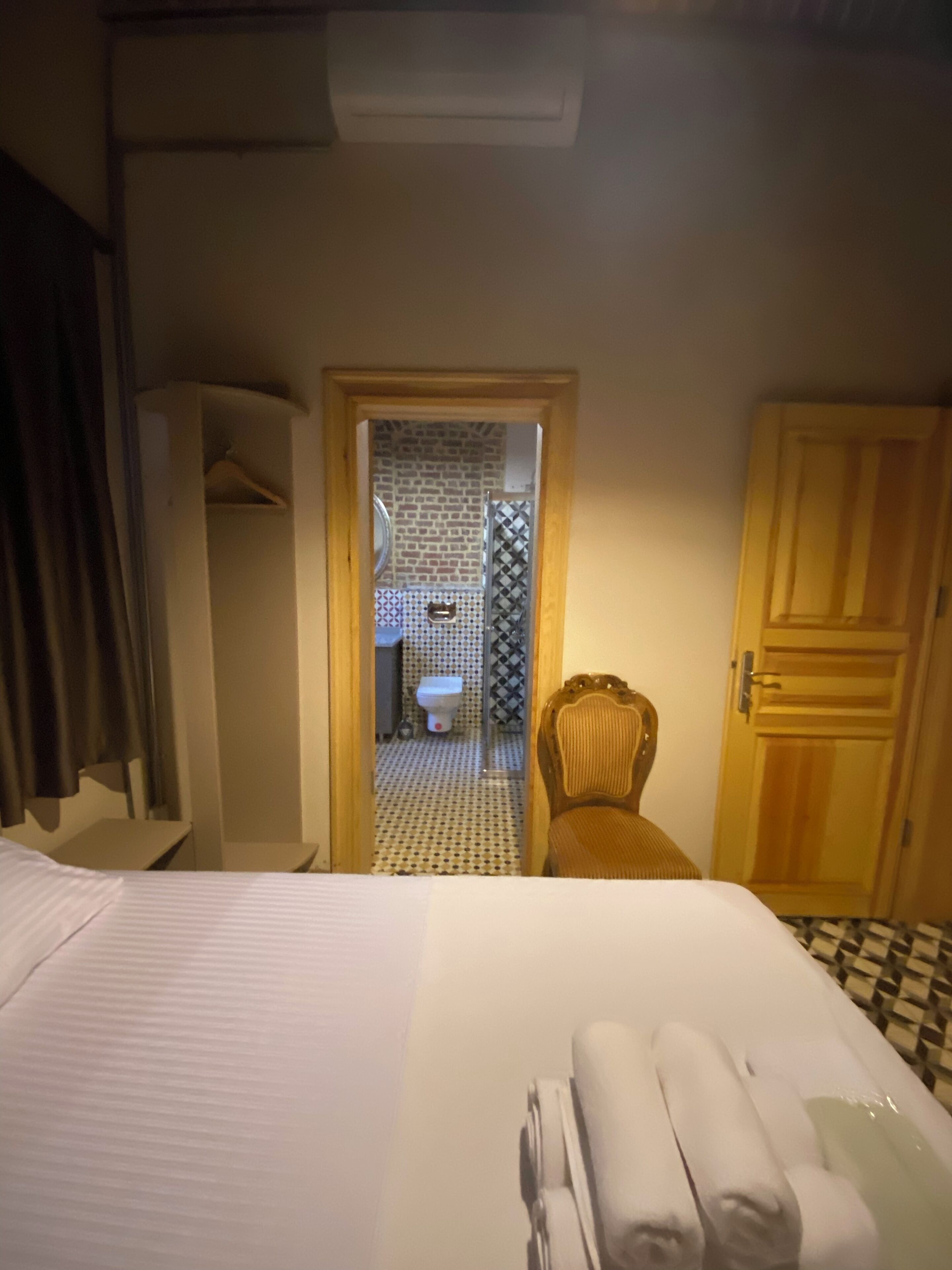 Habitación de hotel con cama, una silla, un armario y un baño visible a través de una puerta abierta.