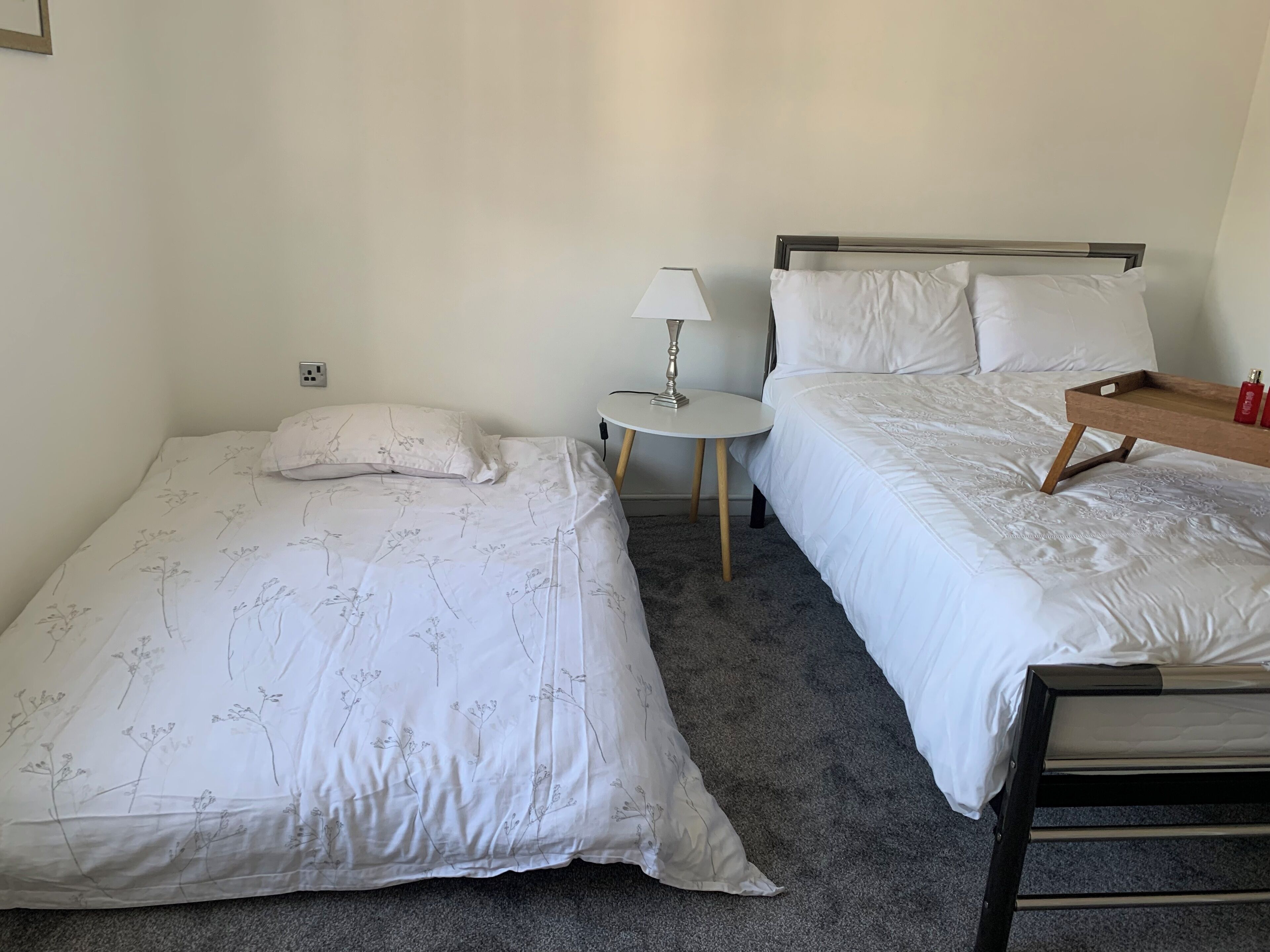 Et lille, rent værelse med to enkeltsenge, et natbord med lampe, og et træsidebord med bakke.
