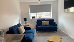 A living room with two blue sofas, a wooden coffee table, a lamp, and a view of a building through a window. - Posh Pad Woolton Village (Liverpool)