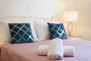 A neatly made bed with a pink bedspread, two blue pillows with gold patterns, and a white towel folded on the bed. - Villa Eleftheria in Spartia (Kefalonia)