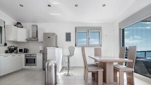 A modern kitchen with white cabinets, a stainless steel range hood, and a built-in oven. There is a dining area with a wooden table and chairs, and a large window offering a view of the sea. - Villa Sapfo in Lefkada (Lefkada)