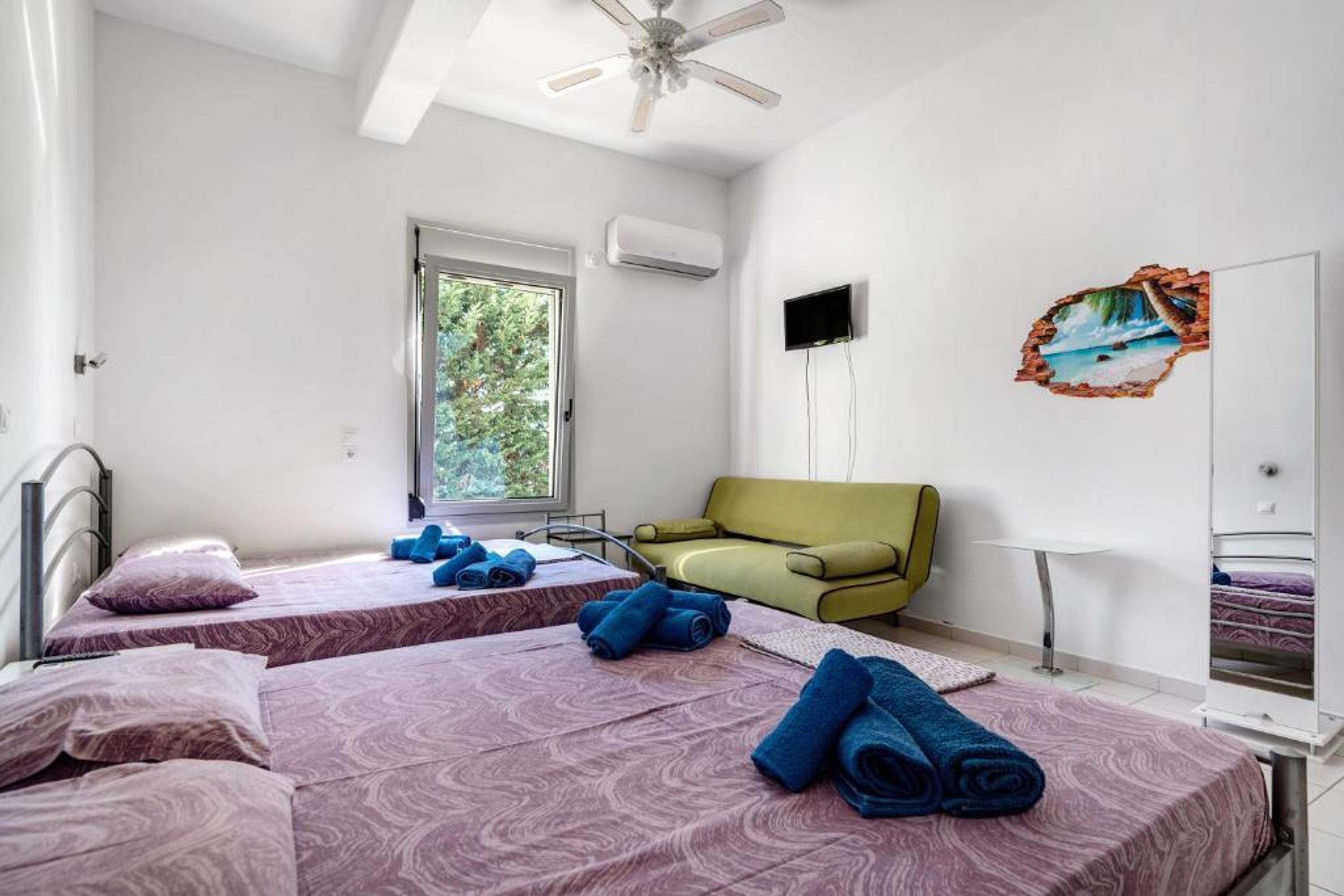 Habitación de hotel con dos camas, un sofá verde, un televisor y una ventana con vistas a la vegetación.