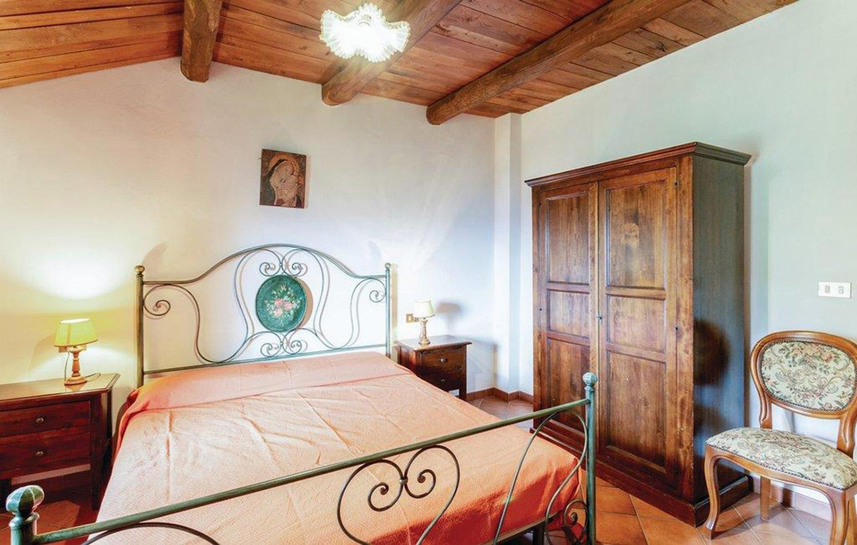 A bedroom with a metal bed frame, a wooden wardrobe, a chair, and a bedside table with a lamp.
