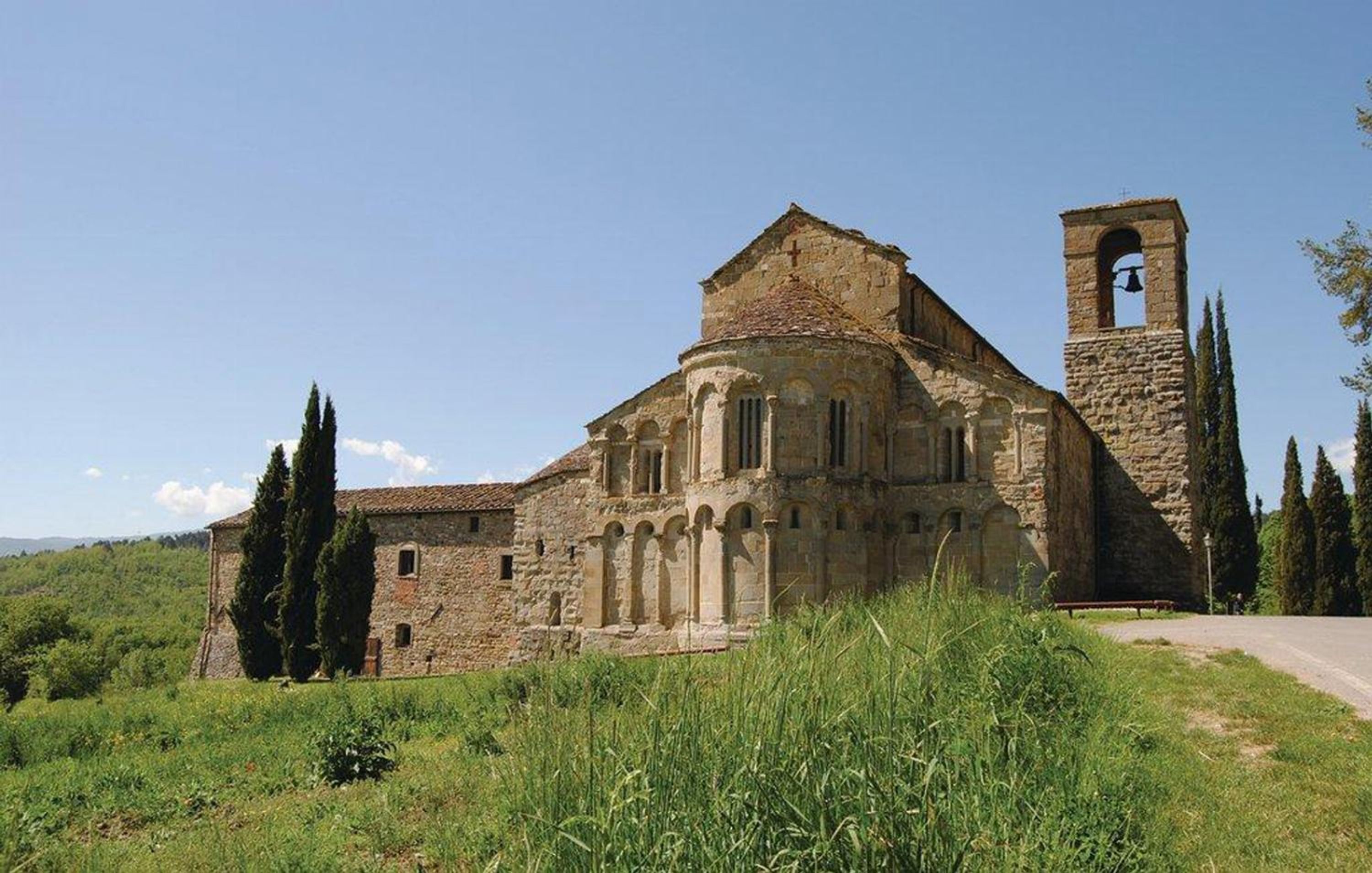 A historic stone building with a bell tower, surrounded by greenery and a clear sky.