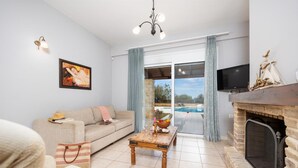 A living room with a sofa, a coffee table, a fireplace, a television, and a view of a pool through a glass door. - Villa Elessa in Rethymno (Rethymno)