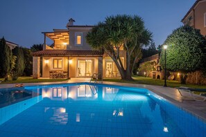 A swimming pool with a tiled deck, a covered outdoor seating area, and a large tree. - Villa Anemoni in Rethymno (Rethymno)