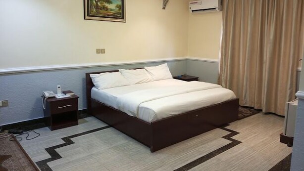 A hotel room with a bed, a bedside table, a phone, a painting on the wall, an air conditioning unit, and a curtain. - Howard Lodgings and Leisure (Calabar)