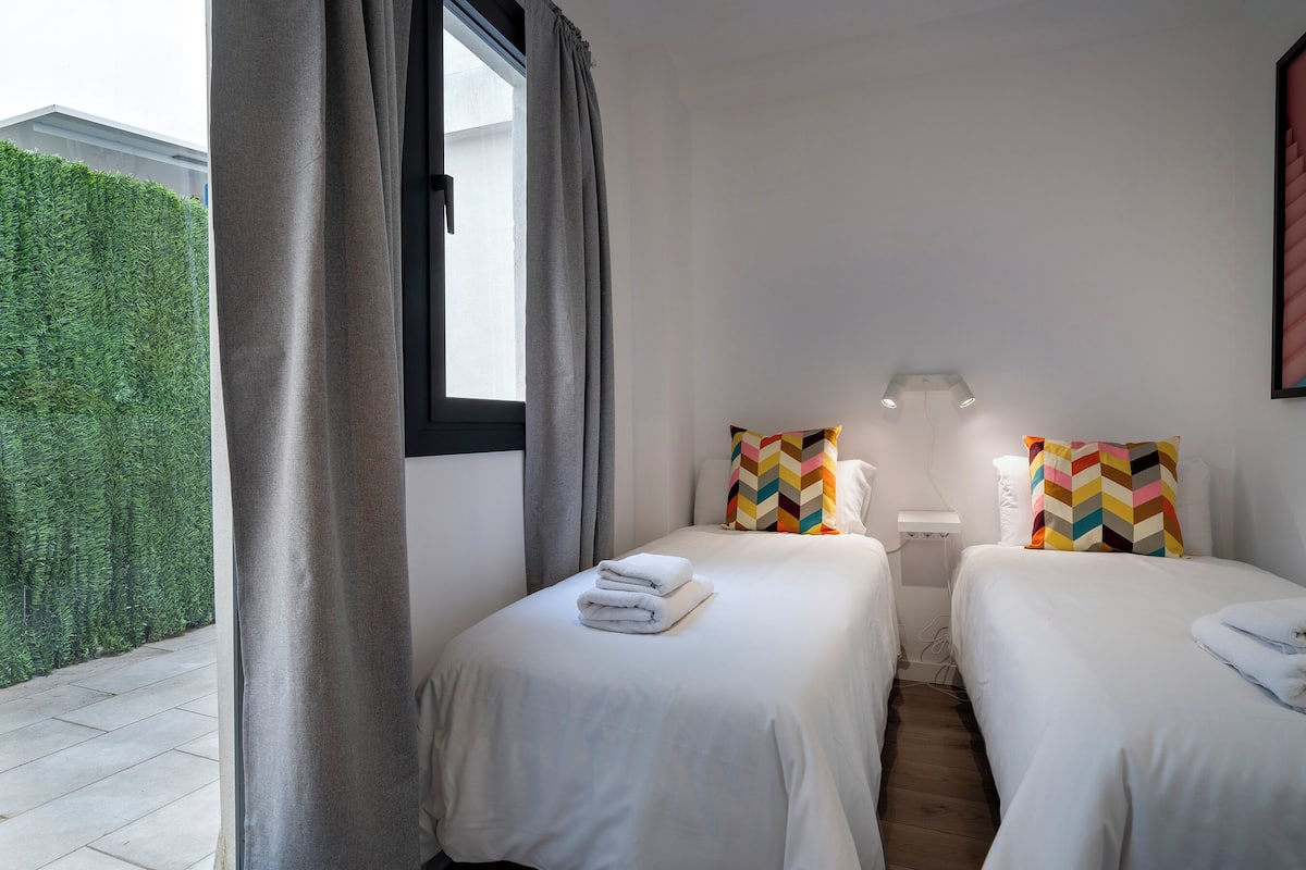 Une chambre d’hôtel avec deux lits, une fenêtre avec des rideaux, une petite table de chevet avec une lampe, et une vue sur une haie verdoyante à l’extérieur.