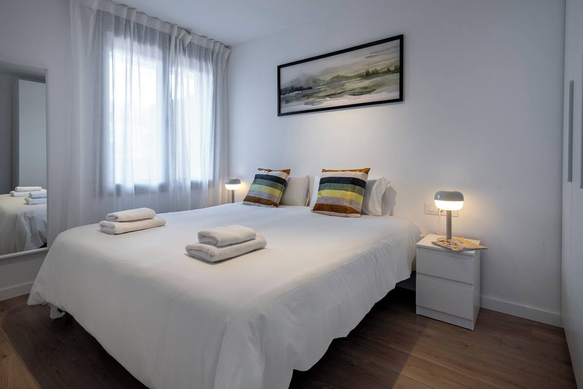 Un lit bien fait, avec du linge de lit blanc et deux oreillers, une table de chevet avec une lampe, et un tableau représentant un paysage, accroché au mur.