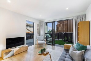 Un salon moderne avec un téléviseur à écran plat, un canapé gris, une table basse en bois et une porte-fenêtre coulissante donnant sur un balcon.