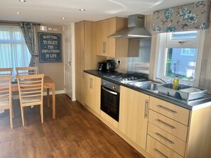 A compact kitchen with wooden cabinets, a black stove, and a window with a view. - 2 Bedroom Self-Catering Chalet near Glasgow City Centre (Stepps)