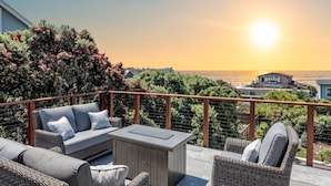 A balcony with wicker furniture, a view of the sunset, and a restaurant in the background. - Miramar Oasis! Ocean View, Hot Tub, Steps2Beach, Roof Deck, Fire Pit, Coastal Trail (El Granada)